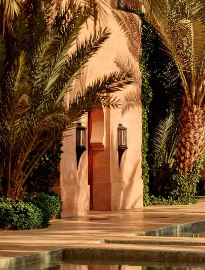 Entrance to a maison at Amanjena resort, Marrakech, with terracotta-toned wooden doors framed by climbing ivy and palm shadows.