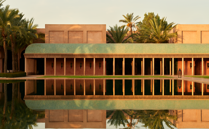Amanjena's central basin with terracotta architecture reflected in still water, surrounded by palm trees.
