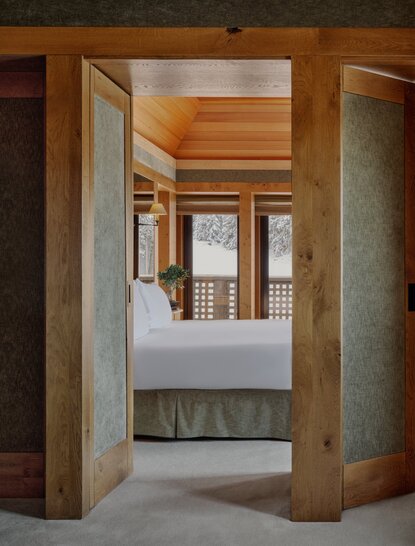 Suite bedroom at Aman Le Mélézin resort, viewed through wooden archway with concrete pillars.
