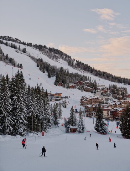 Skiers and snowboarders on snowy slopes at Aman Le Mélézin resort, with forested mountainside and alpine village visible in background.
