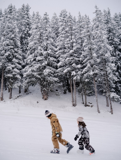 Skier and snowboarder on snowy slope at Aman Le Mélézin resort, surrounded by snow-covered pine forest.