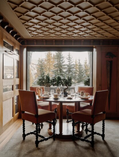 Dining area at Aman Le Mélézin with round table, wooden chairs, and forest views through large windows.
