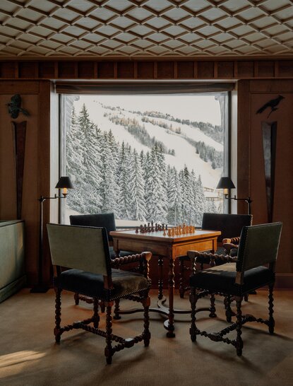 Salon at Aman Le Mélézin resort with seated chairs and table overlooking snowy Alpine landscape through large window.