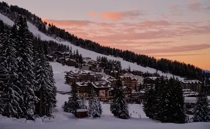 Exterior view of Aman Le Mélézin resort at sunset, with snow-covered slopes and forested mountains under a pink-hued sky.