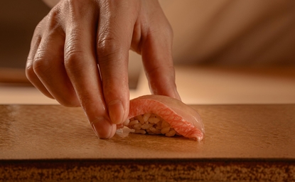 Hand placing sesame-topped appetiser on wooden surface at Aman Nai Lert Bangkok dining venue.