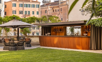 Aman Venice bar area with wooden counter and seating on manicured lawn, Venetian architecture beyond.