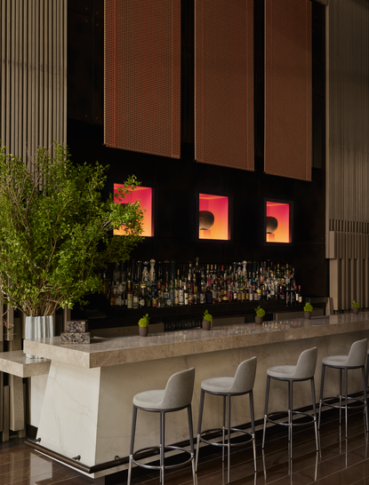 Aman New York's lounge bar features a minimalist bar counter with neutral stools beneath illuminated shelving displaying spirits and glassware.