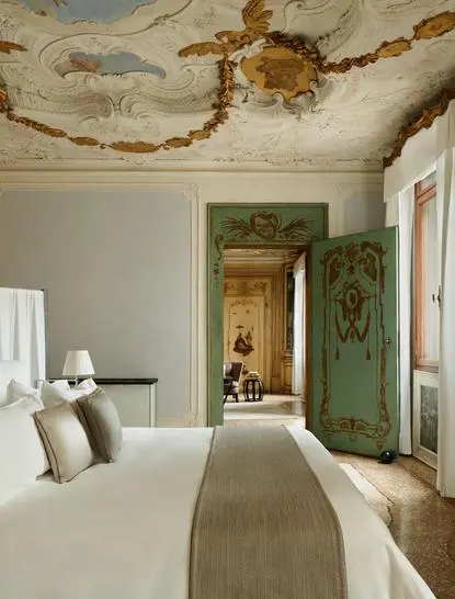 Alcova Tiepolo Suite bedroom at Aman Venice, with frescoed ceiling, green door, and neutral furnishings.