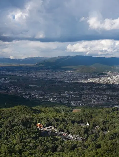 Wenfeng Temple nestled in verdant hills above the Amandayan valley, with dramatic clouds gathering over distant mountains.