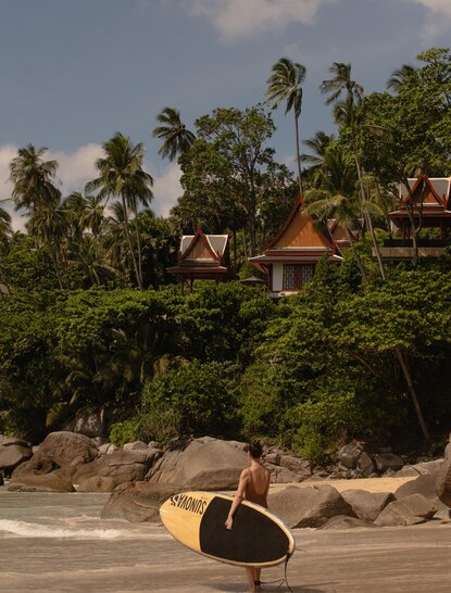 Frau auf Stand-up-Paddleboard vor Strand mit Palmen und Resort-Gebäuden, Amanpuri, Thailand.