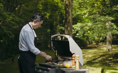 Chef prepares food at an outdoor barbecue surrounded by lush greenery at Aman Kyoto resort.