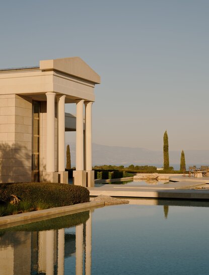 Amanzoe resort at dawn, with stone pavilion reflected in still water and distant monument visible across the landscape.