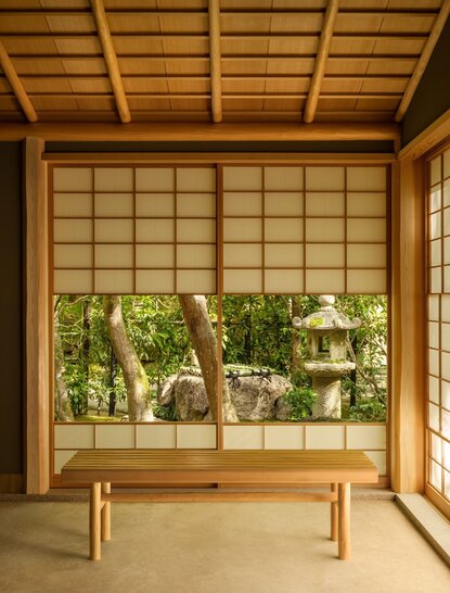 安缦京都度假村的茶室内部，木制格子窗框前放置一张简约木凳，窗外可见绿植。
