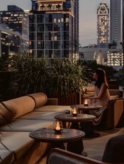 Rooftop seating area at Aman Nai Lert Bangkok with city views at dusk, featuring contemporary furniture and potted plants.