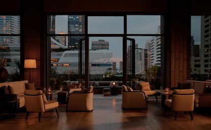 Terrace dining area at Aman Nai Lert Bangkok with floor-to-ceiling windows overlooking the city skyline at dusk.