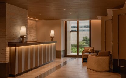 Warm wooden wellness clinic reception at Aman Nai Lert Bangkok, with desk and seating area overlooking gardens.