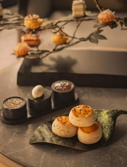 Afternoon tea service at Aman Nai Lert Bangkok with pastries, cocktails and floral arrangements on slate table.