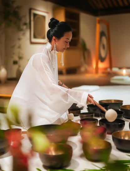 Woman in white shirt preparing tea at Amanfayun, with traditional tea vessels and warm candlelit setting.