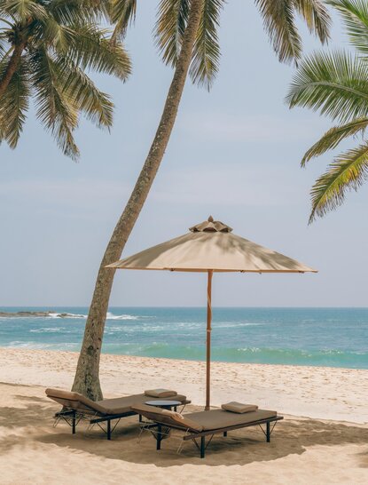 Wooden sunbeds and parasol beneath palm trees on Amanwella's sandy beach, with turquoise waters beyond.