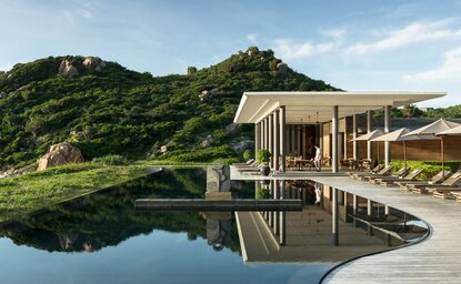 Amanoi beach club with wooden pavilion reflecting in still water, verdant hillside beyond.
