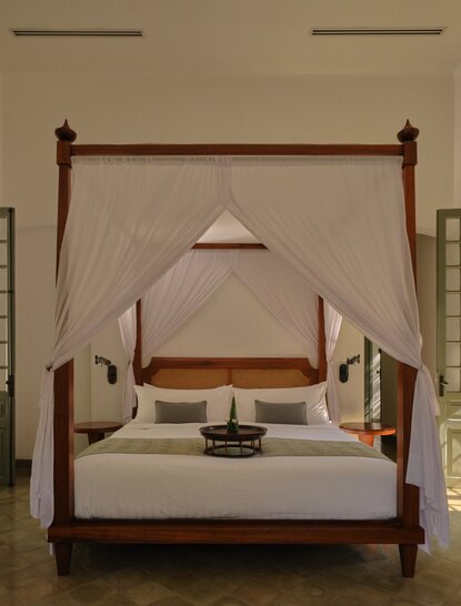 Amantaka bedroom with four-poster bed draped in sheer fabric, wooden furnishings and neutral tones.