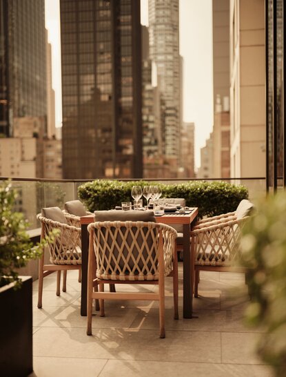 Terrazza dell'hotel Aman New York con tavolo e sedia in vimini, circondata da piante verdi e grattacieli sullo sfondo.