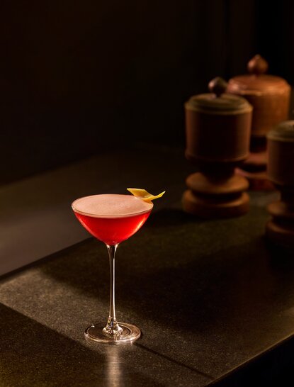 Amanoi bar cocktail in a coupe glass with red liquid, shaker and jigger in background.