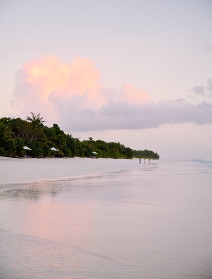 Amanpulo's shoreline at dusk, with soft pink clouds reflected in calm waters and forested peninsula beyond.