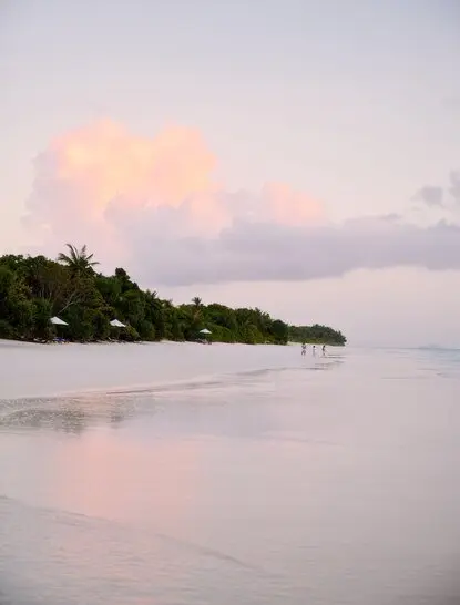 Amanpulo resort beach at dusk, soft pink sky reflecting on calm water, tree-lined shore in distance.