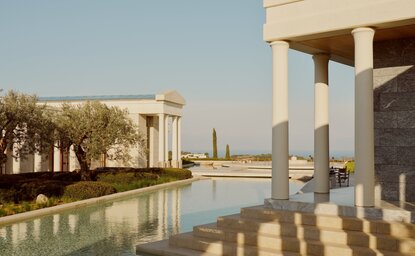 Waterfront view of Amanzoe resort with modern architecture and reflecting pool against clear Mediterranean sky.
