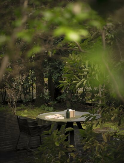 A solitary wooden table and bench set among lush greenery at Aman Kyoto, dappled with soft natural light filtering through the trees.