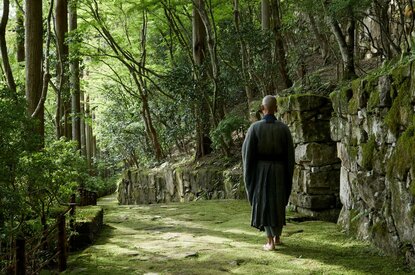 A figure in dark clothing walks along a peaceful stone pathway lined with moss-covered lanterns and tall trees at Aman Kyoto resort.