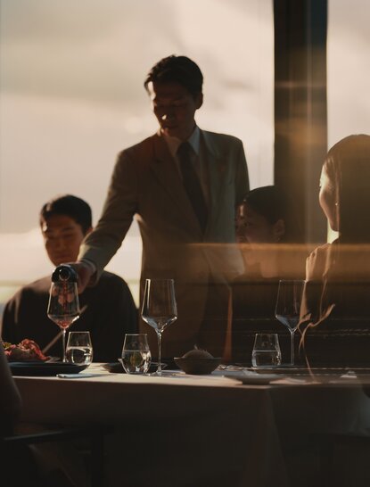Adult and child dining together at Aman Tokyo hotel, with Tokyo skyline visible through windows behind them.