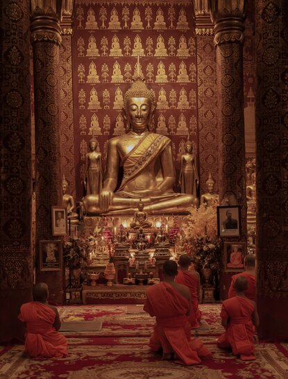Monks in crimson robes meditate before a gilded Buddha statue at Amantaka, Laos.