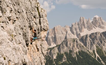 Rock climber ascending a dramatic limestone cliff face in the Dolomites, Italy, with jagged peaks visible beyond.