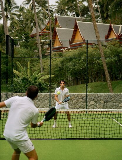 Padel player hitting ball during match at Amanpuri, Thailand, with tropical greenery beyond the court.