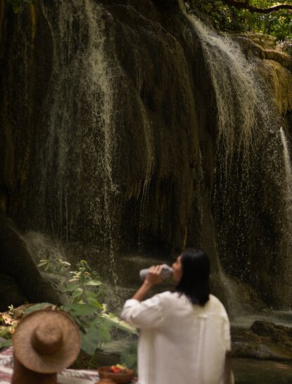 A guest in white clothing stands before a cascading waterfall at Amanwana, Indonesia, with a straw hat visible in the foreground.