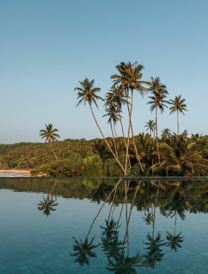 Amanwella's infinity pool overlooking tranquil waters with palm trees reflected below.