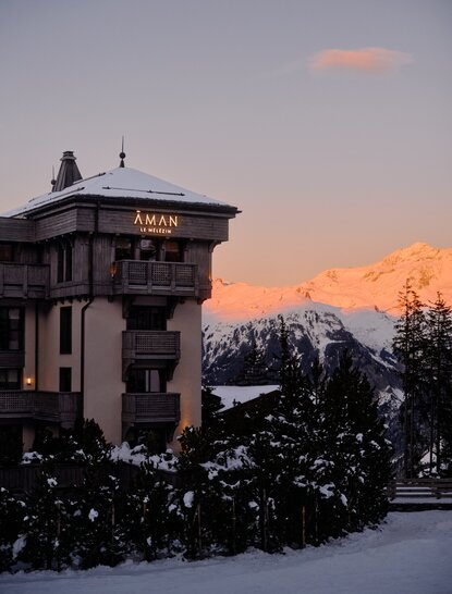 Aman Le Mélézin exterior at dusk, snow-covered grounds with Alpine peaks beyond.