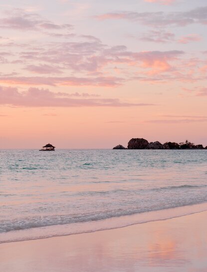 Sunset view from the beach at Amanpulo, Philippines, with soft pink and orange light reflecting on the sea and sand.