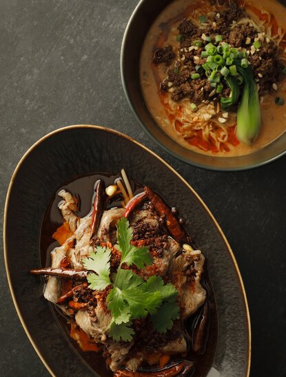 Overhead view of two dishes at Aman Tokyo: a bowl of spiced broth with vegetables, and a plate of stir-fried meat with fresh herbs.