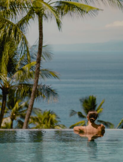 Baigneur dans la piscine à débordement d'Amankila avec vue sur l'océan et palmiers, Bali.