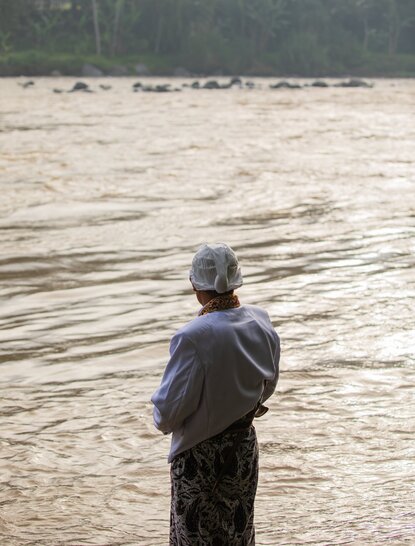 Solitary figure in dark robes standing at water's edge during Ruwatan wellness ritual at Amanjiwo.
