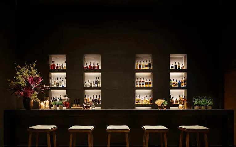 养云安缦度假村的餐厅酒吧，夜晚灯光照亮展示柜内的酒瓶，木制吧台前方摆放座椅。