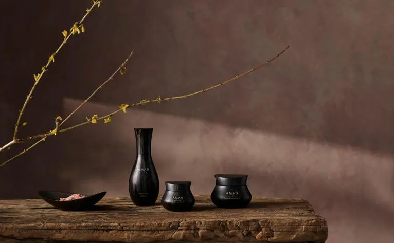 Aman Skincare campaign shot featuring dark glass bottles and vessels arranged on a wooden surface with dried branches in an office setting.