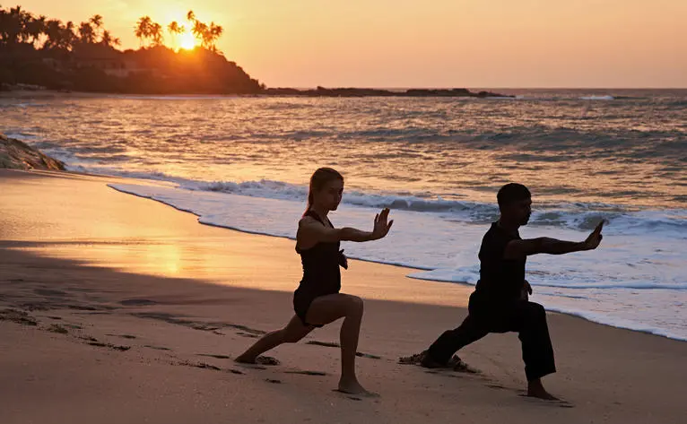 Two people practising yoga on Amanwella's beach at sunrise, with golden light reflecting on the sand and sea.