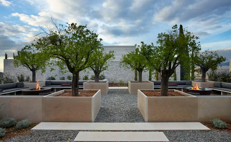 Symmetrical dining villa courtyard at Amanzoe with tree-lined pathways and stone seating areas under blue sky.