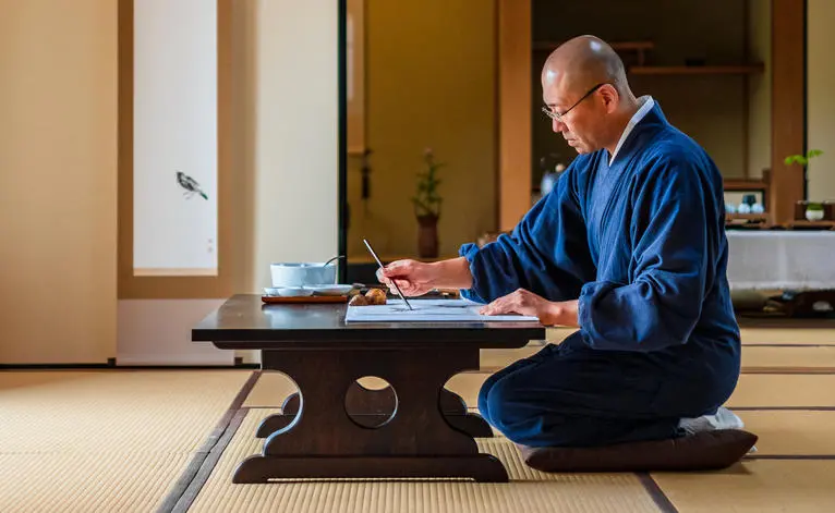 Man in blue robe seated on tatami mat at low desk, practising ink painting at Aman Kyoto resort.