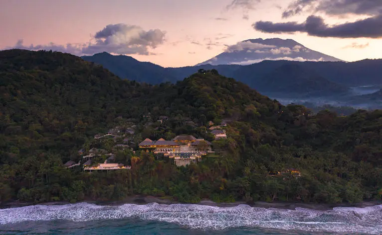 Amankila resort aerial view at dusk, with terraced architecture nestled among forested hillsides overlooking the coast of Bali, Indonesia.