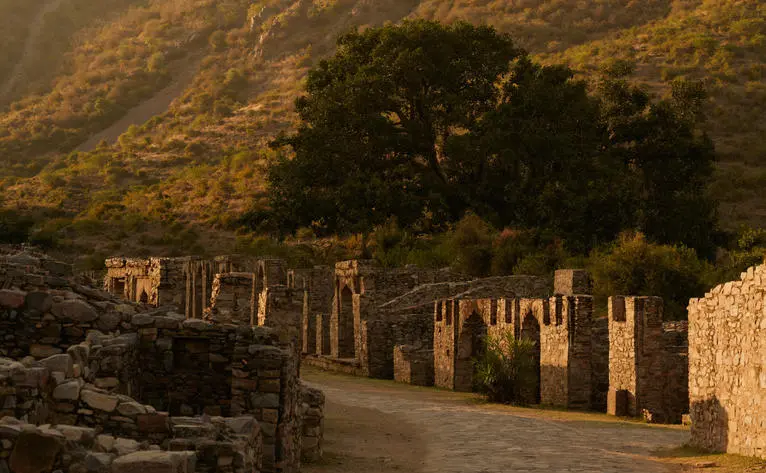Gateway and tree at Amanbagh, with village ruins and hills beyond in golden evening light.